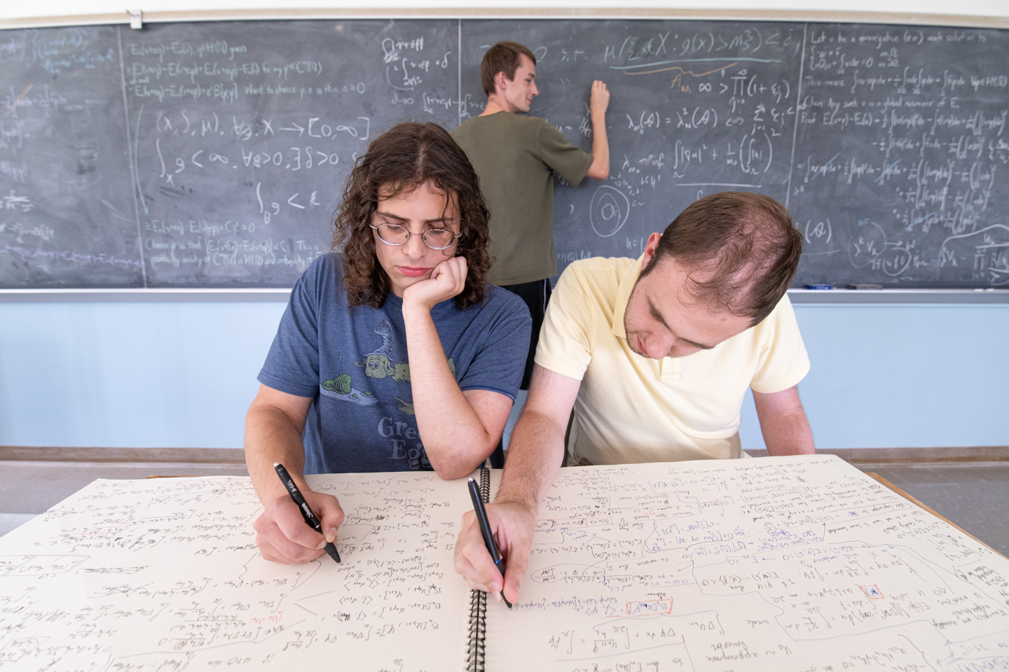 Two students in the foreground work on equations on a large notepad while a third student in the background works on equations on a blackboard behind them. 