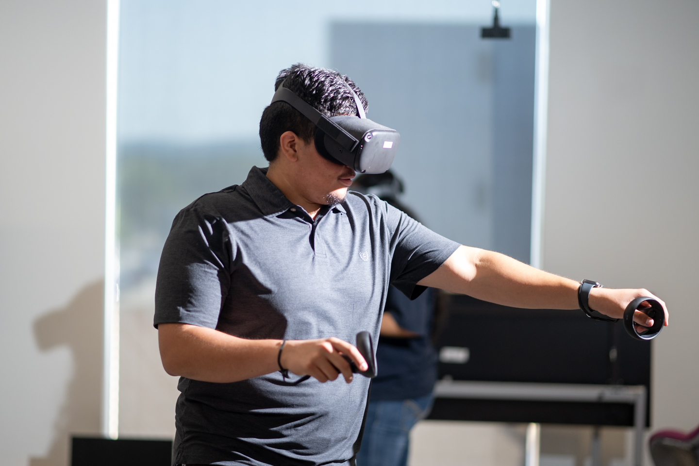 A student wearing a VR headset is in the center of the frame in a modern space. He is facing towards the left side of the frame and his left arm is fully extended. His right arm is lifted half way and bent at the elbow. 