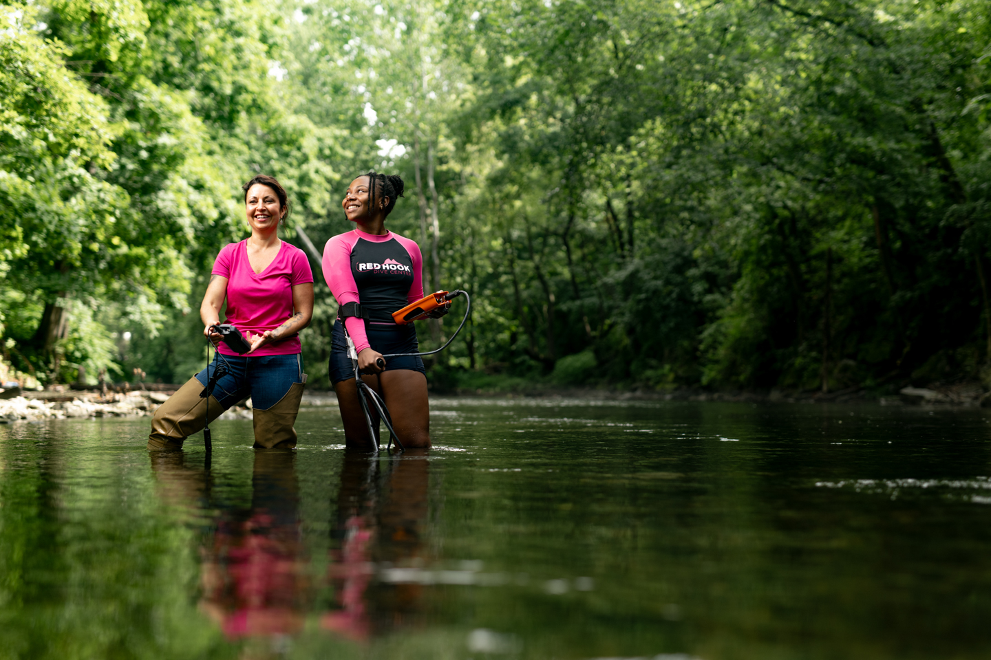 Two women dressed in pink stand in a knee deep creek. They are both holding research equipment. Trees and green leaves can be seen all through the background of the image. 