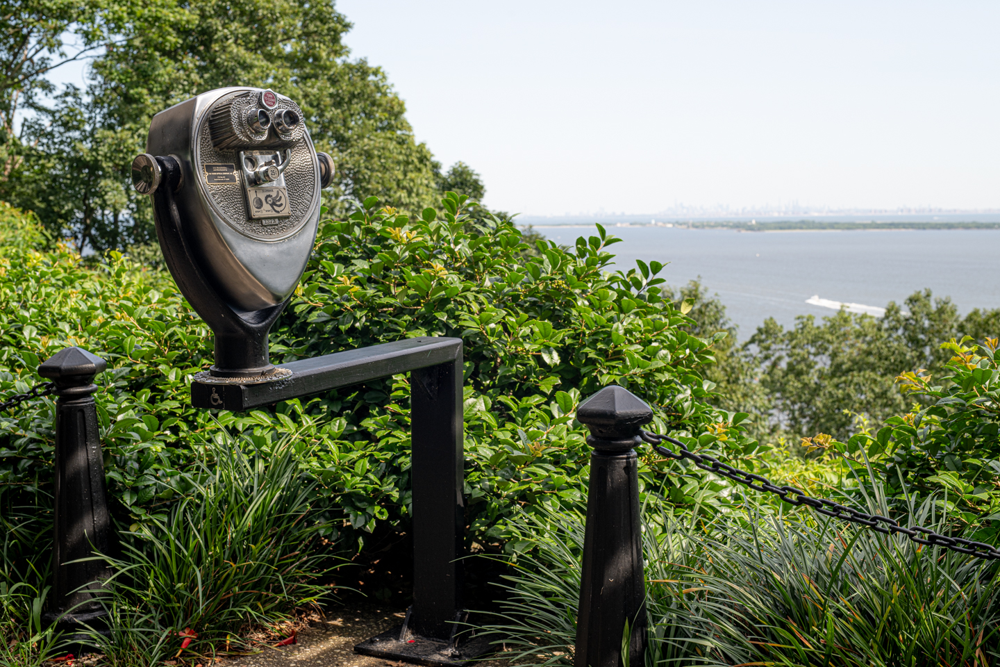 A pair of timed binoculars is mounted on a scenic overlook, on the left side of the frame set against green leafy growth, of the Atlantic Highlands National Park in Sandy Hook NJ. In the distance The NYC skyline is visible. 