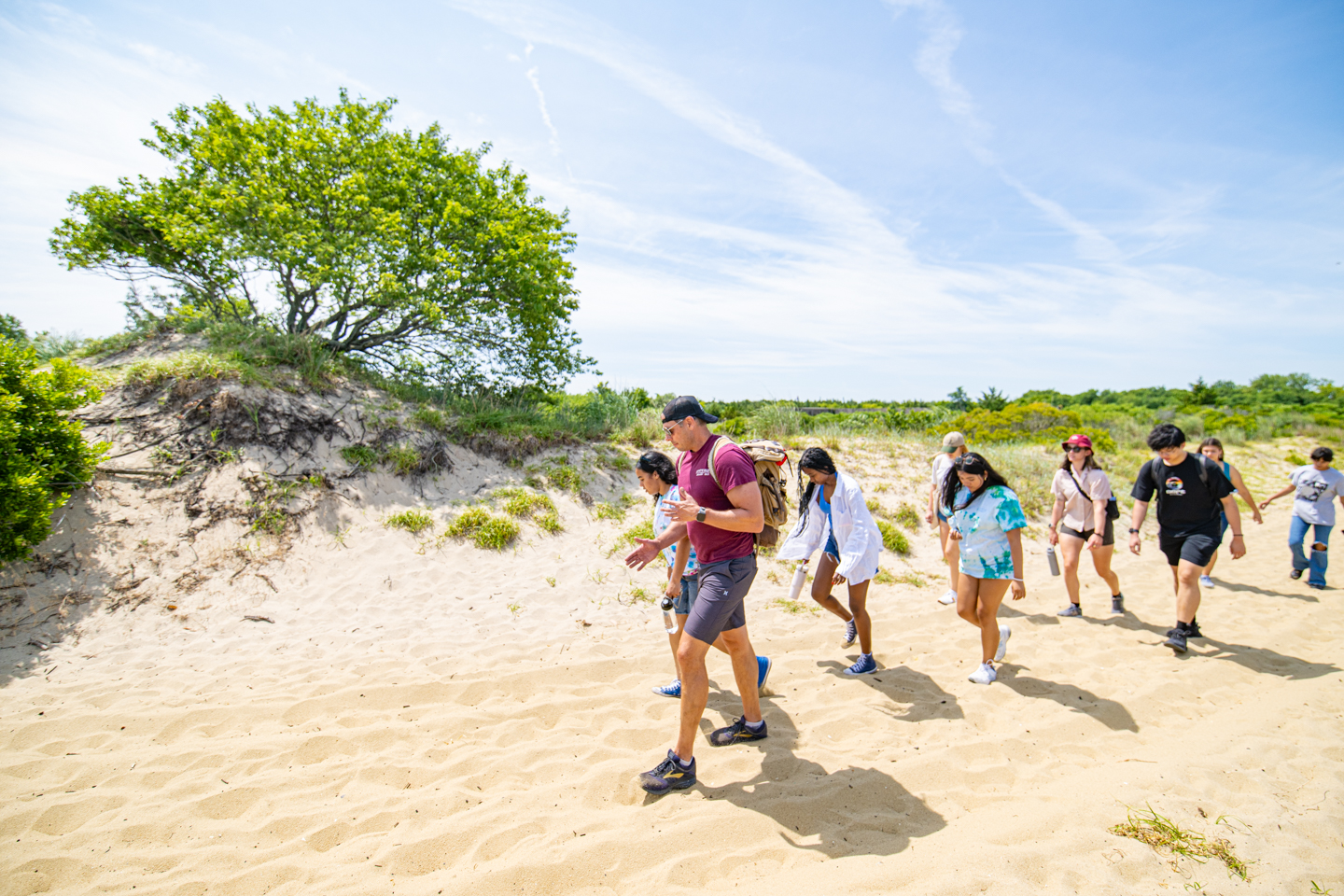 A professor leads a group of students through a sandy beach path. Green Grass and small tree are on the left side of the frame. 