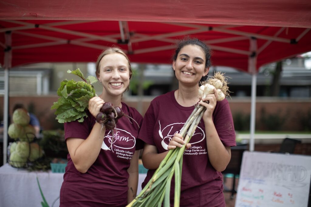 Two students hold up leeks and beets under the red LaFarmstand tent.