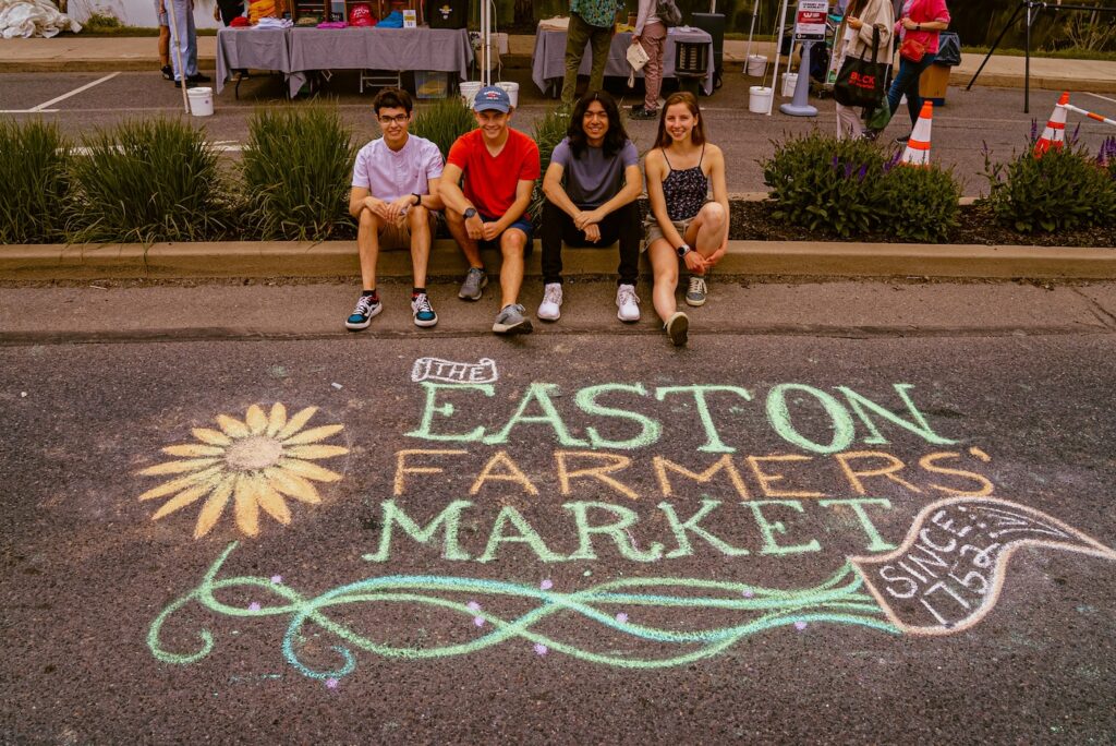 4 students sit on the sidewalk in front of a chalk design that reads Easton Farmers' Market.