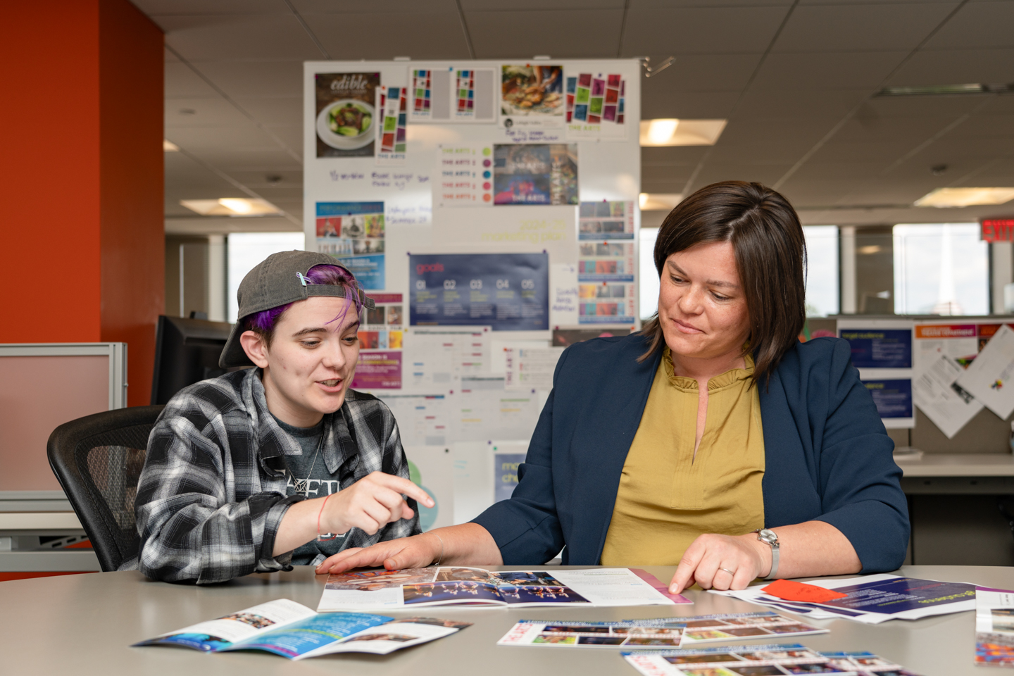 A student on the left side of the frame, interns with a mentor on the right side of the frame. In the background a bulletin board with their work hung on it can be seen. 