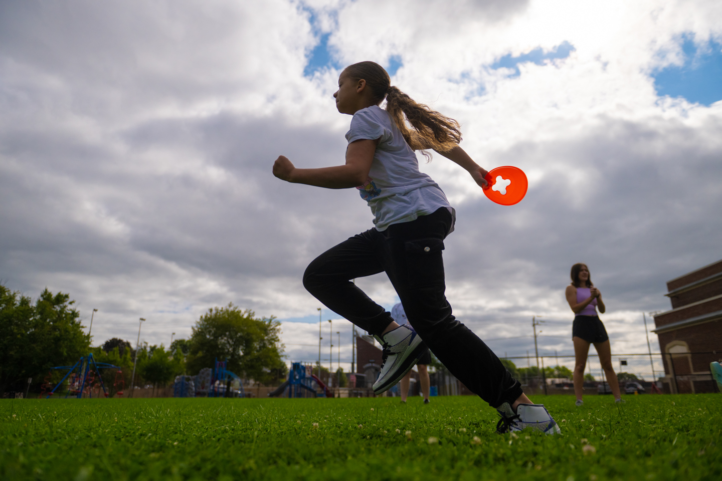 A young girl holding a small cone sprints across the frame from right to left. 