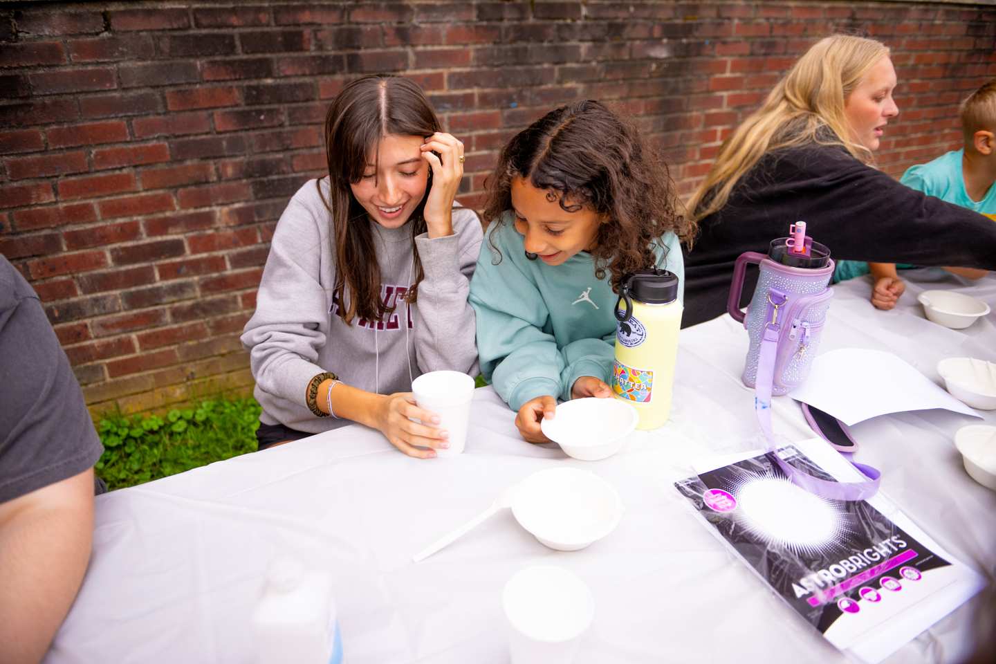 A young woman and little girl work together to make "elephant toothpaste." They are working at a picnic table with dropcloths on it, and a brick wall is directly behind them. 