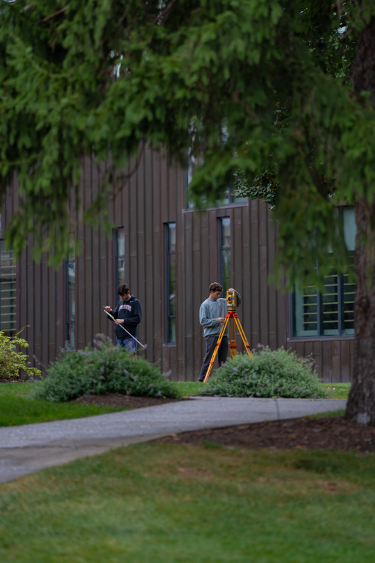 Two people in the distance using land surveying equipment framed by an evergreen tree.