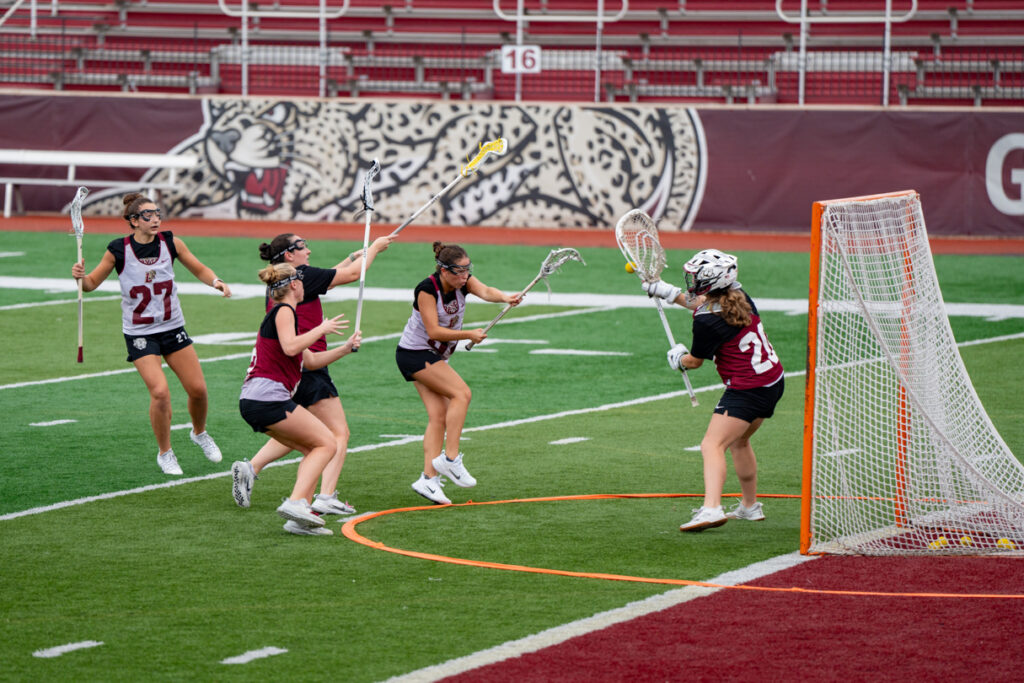 A lacrosse player shoots the ball at the goalie while defenders surround them.