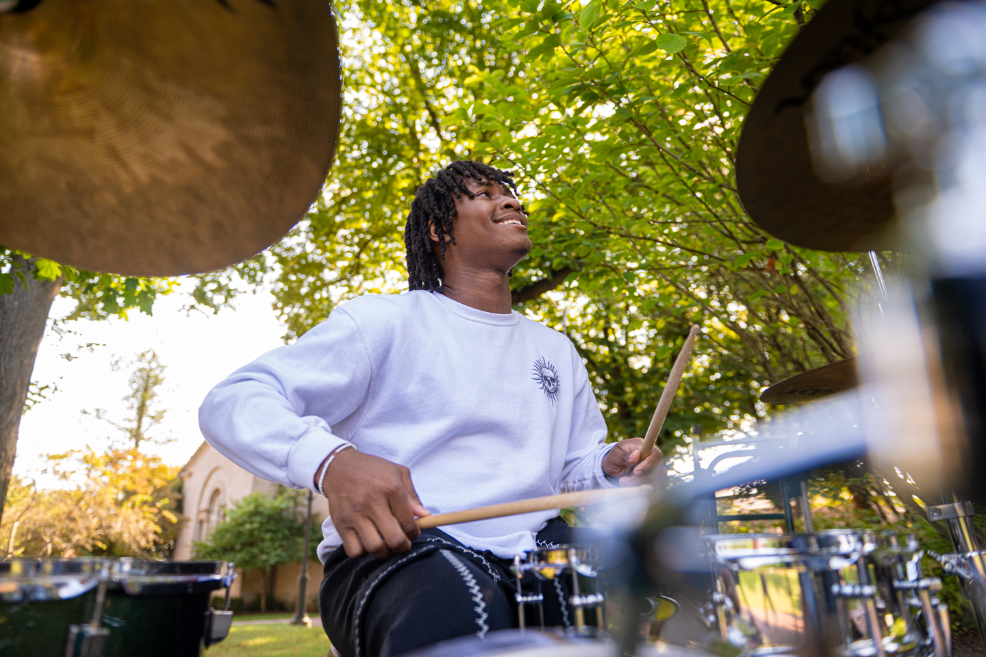 A young person is seated in the center of the frame at a black drumset with chrome trim. Visible in the top left and right of the frame are two cymbals. The young person is holding drumsets and appears to be playing the drums. 