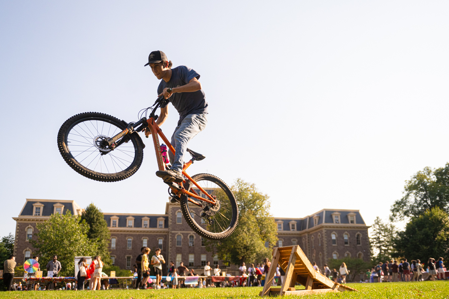 A young man jumps a mountain bike off of a small ramp in the foreground. He is wearing a cap, gray t-shirt and light blue jeans. In the background there is a crowd of people attending a tabling fair. Further in the background is Pardee Hall. 