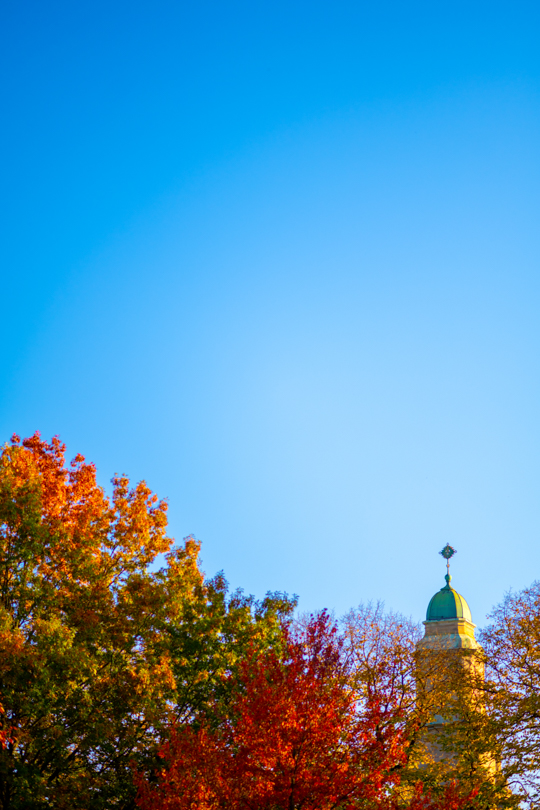 The top of Colton Chapel peaking through red and orange leaves in front of a bright blue sky.