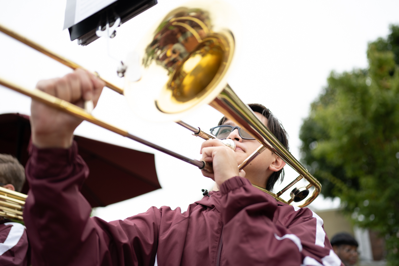 A student dressed in a maroon windbreaker plays a trombone. The viewpoint of the camera is lower looking up. the student is in the right side of the frame facing left. 