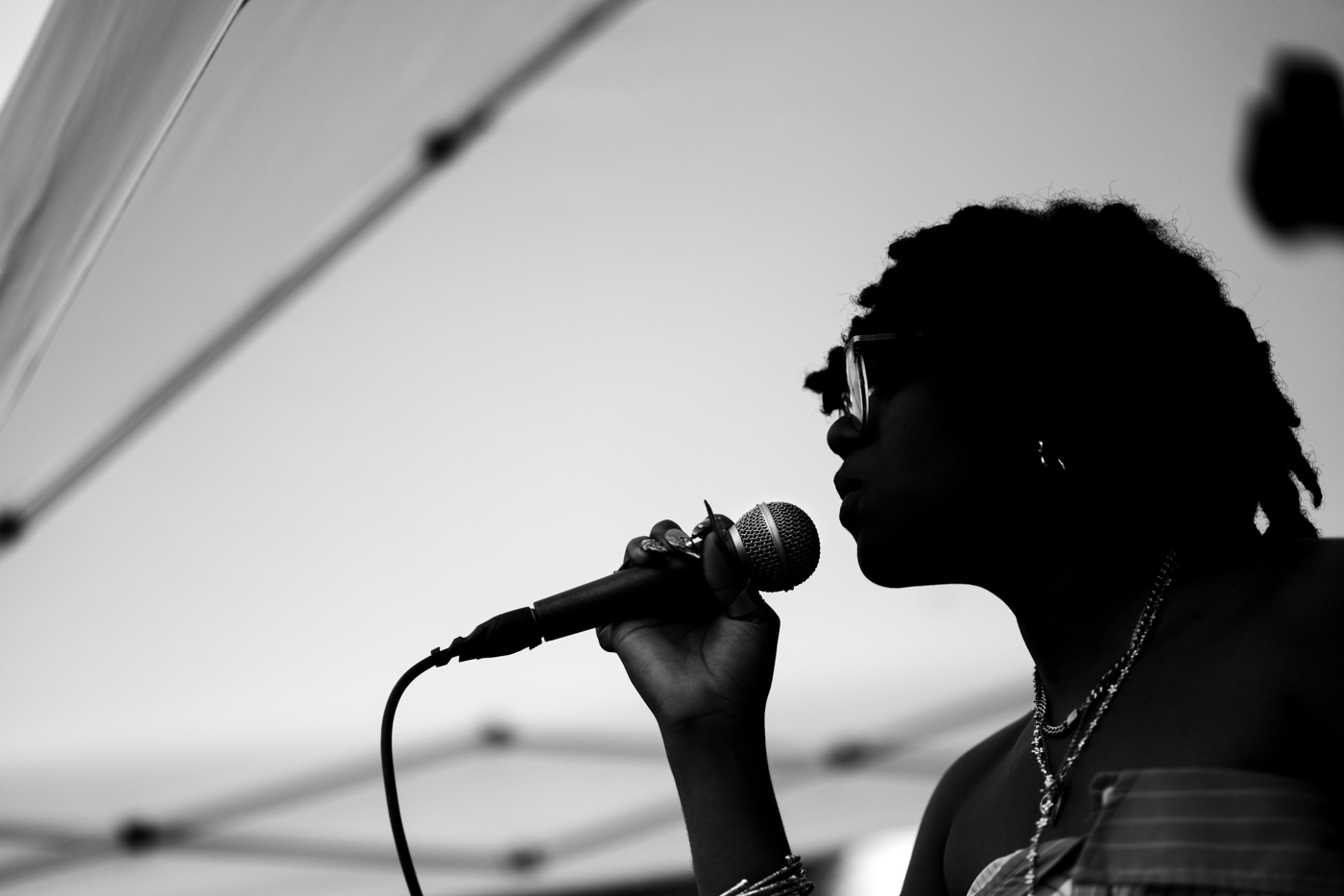 This black and white photo depicts the silouhette of a woman singing into a microphone, set against the white surface of an outdoor events tent. 