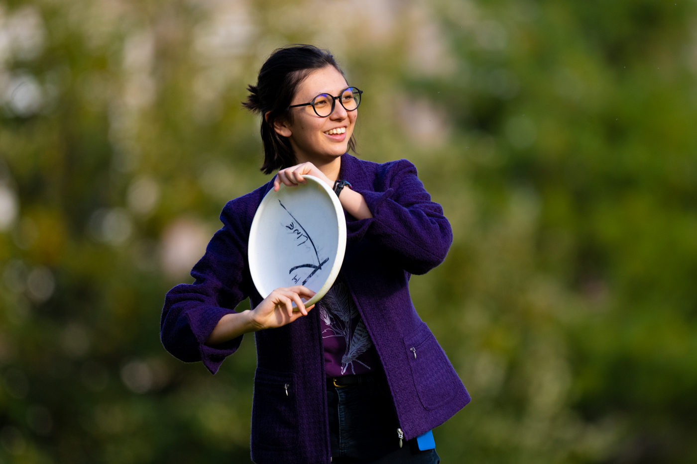 A college student with dark long hair and a purple shirt is seen holding a frisbee she is about to throw. Green trees can be seen blurry in the background. 