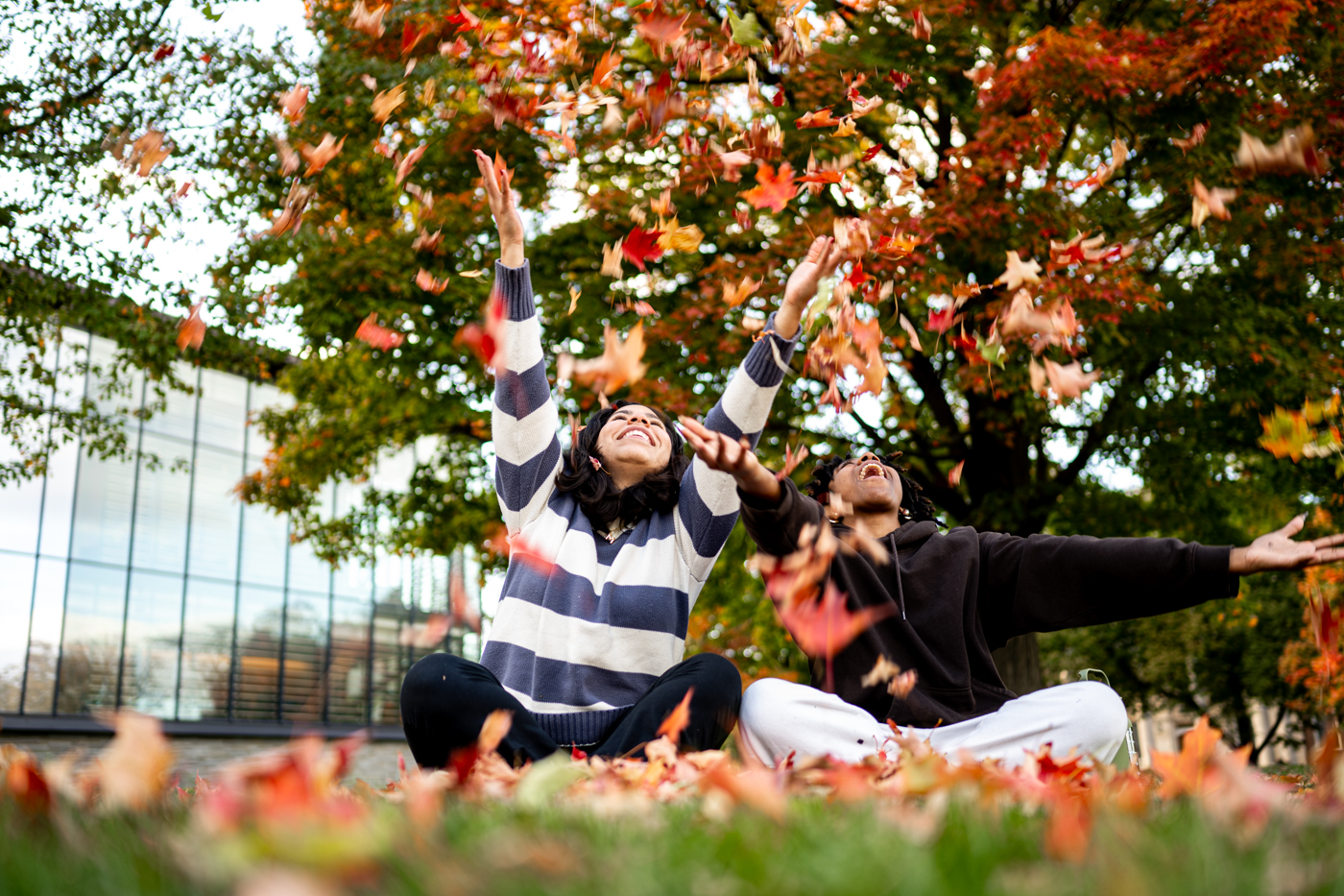 two people are sat next to each other with their legs crossed. They are both tossing leaves above their head. They are seated in front of a large tree. The leaves are in the midst of changing color from green to orange. 