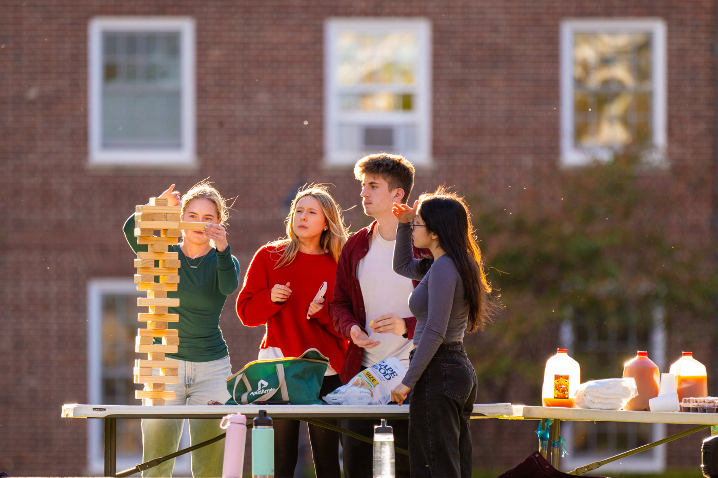 A group of students can be seen playing a large set of Jenga. nex to them on a table gallon jugs of apple cider can be seen. 