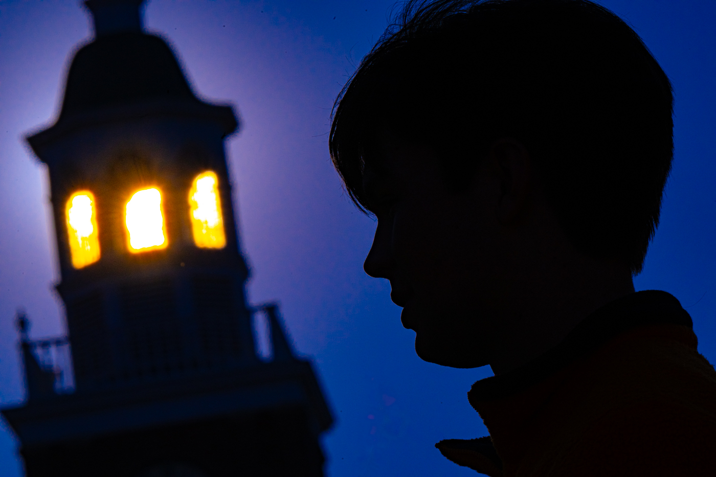 the silhouette of a cupola and the profile of a college aged male are backlit by the sun and set against a clear blue sky. 