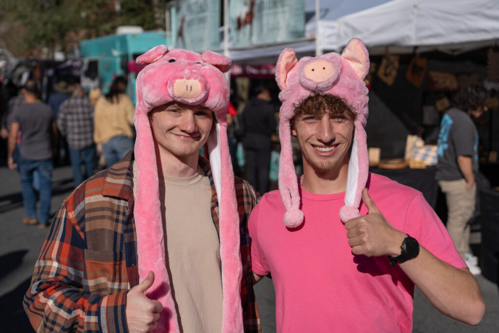 2 people wearing fuzzy pig shaped hats smile and give a thumps up.