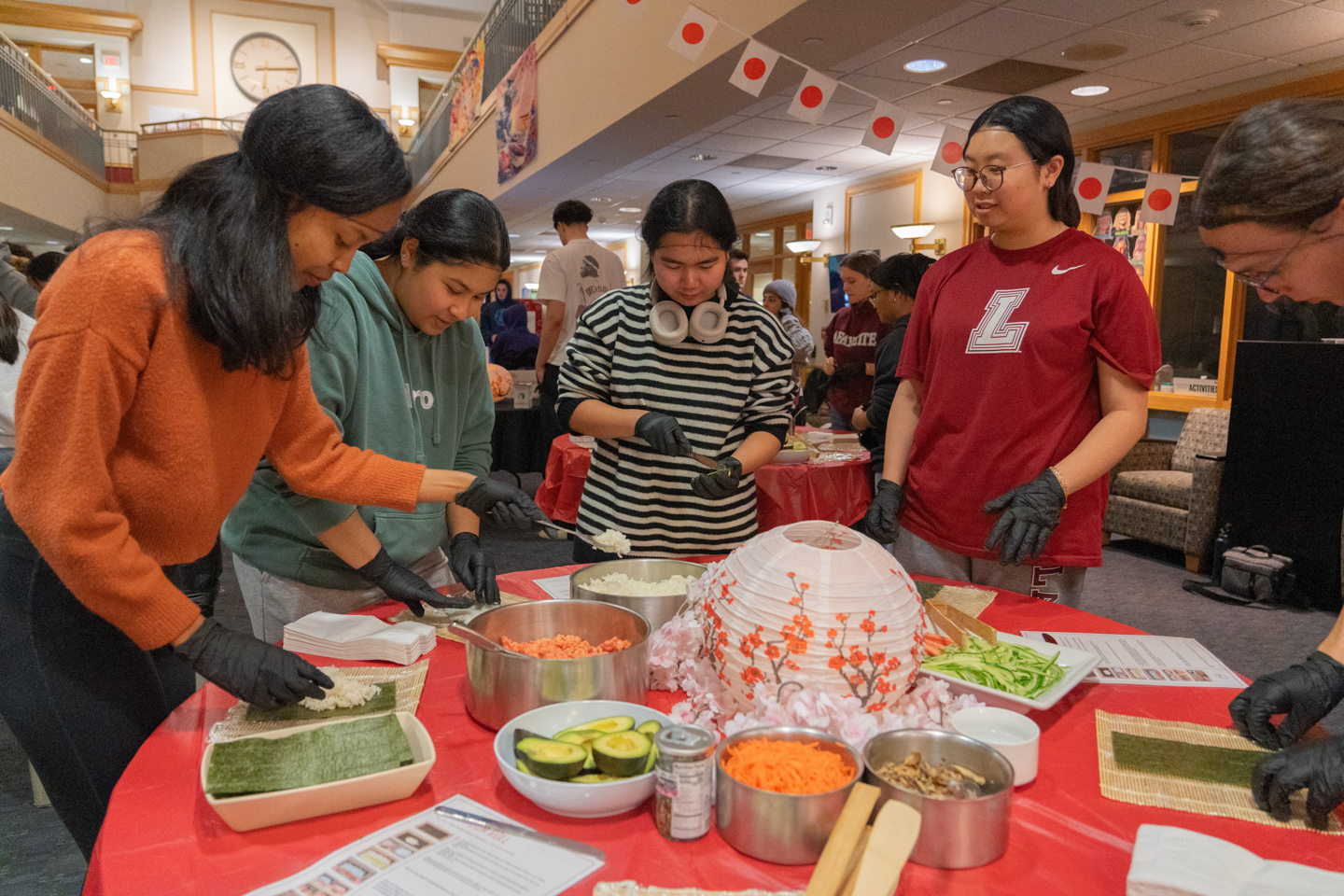 Four students stand next to each other around a round red table. The table is covered with many various types of ingredients used to make sushi. 