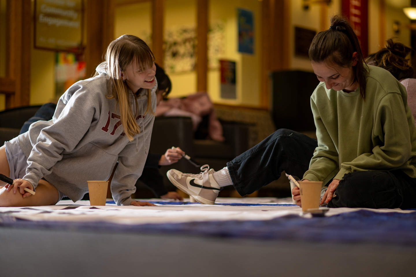 Two students are seated on the floor as they paint rivalry slogans onto white banners.