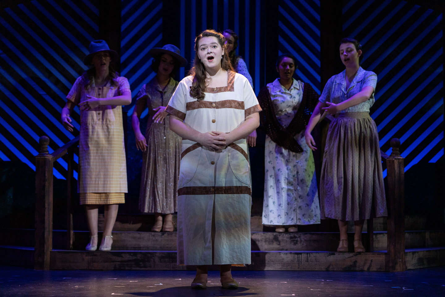 five student peformers can be seen on stage. The one in the center is lit with a spotlight. The background s lit with blue lights. 