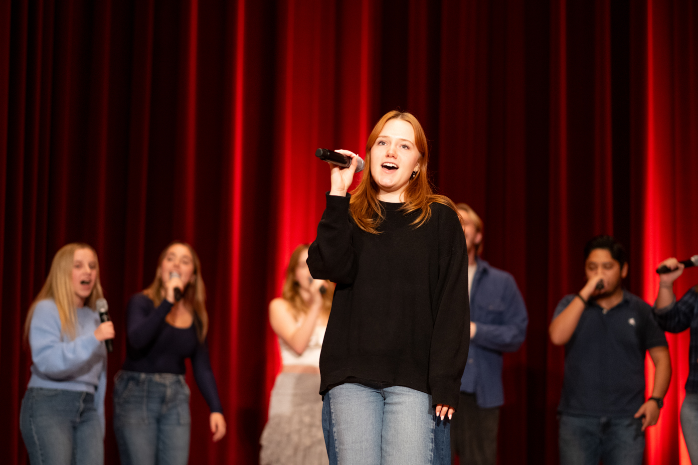 The lead singer in an a capella group takes center stage. There is an uplit red curtain behind her and the group. 