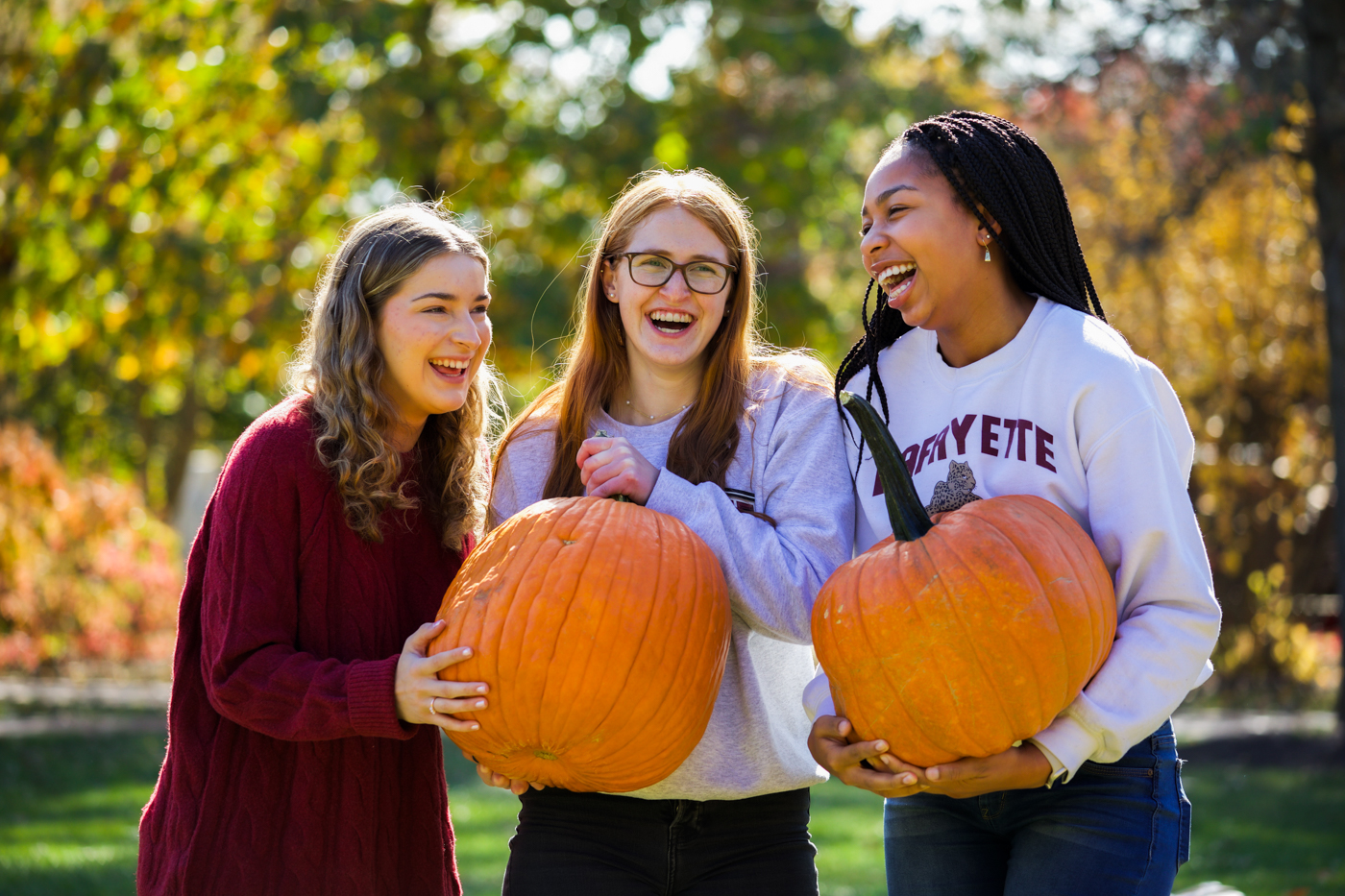 Three female college students in sweaters are standing next to each other facing the camera. One in the middle and one on the right are holding a pumpkin. They are all laughing. Behind them, trees in various states of change can be seen out of focus. 