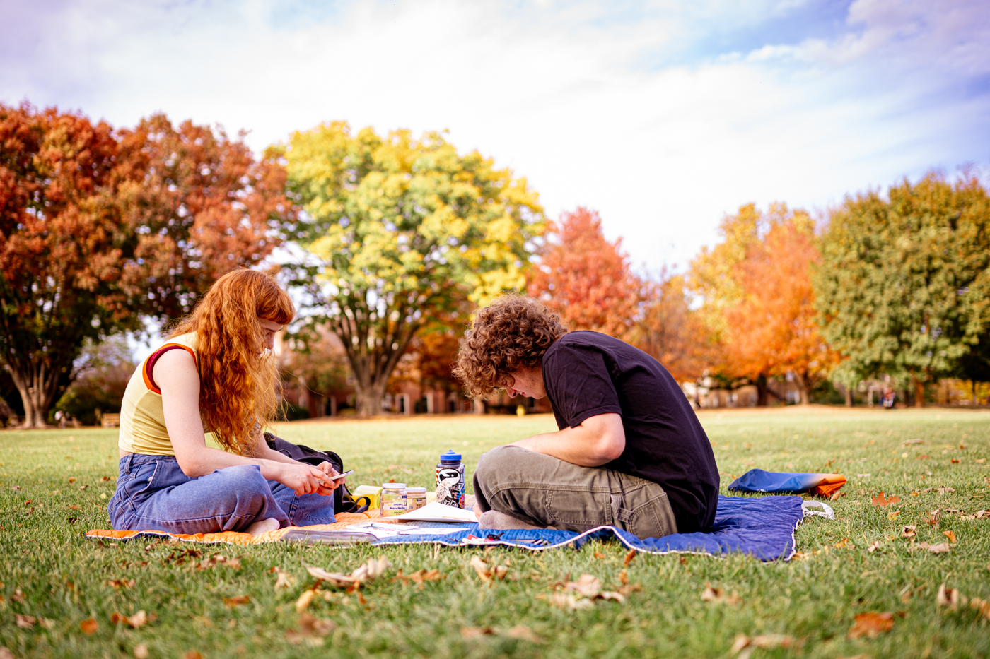 a male and a female student sit across from each other on a blanket,on a sunny fall day. Behind them are trees in various states of change in the fall season. 