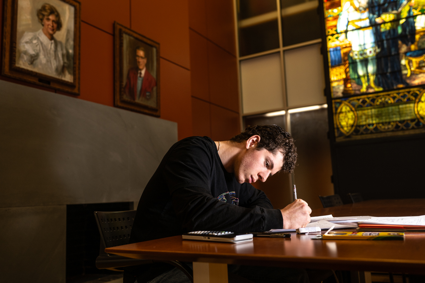 A college aged student is seated and leaning over a table in a wood paneled room with, preserved, stained glass window displays. 