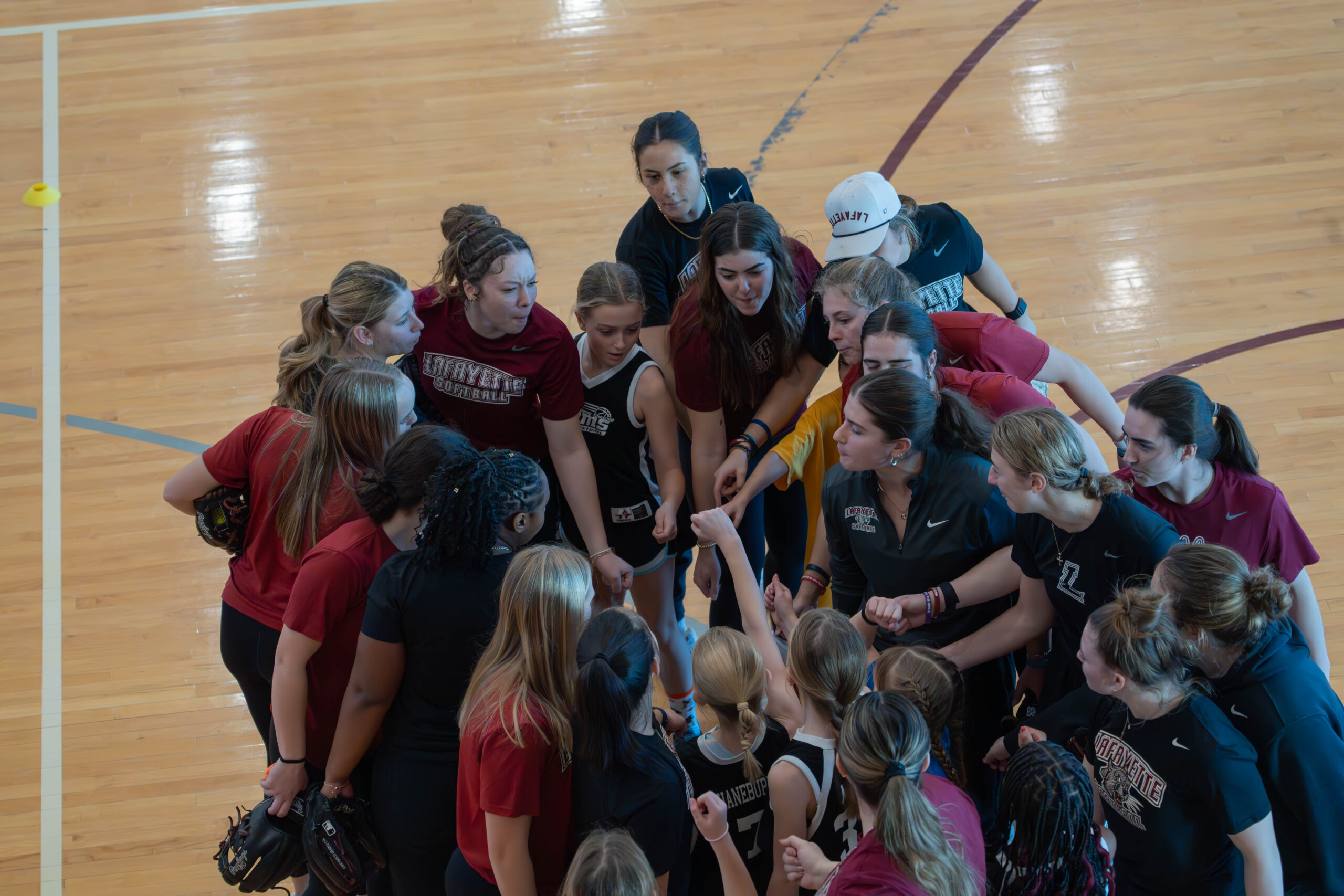 A group of collegiate women athletes and elementary school girls participate in an all hands in huddle. The photo is taken from above. They are in a gymnasium with basketball court markings. 