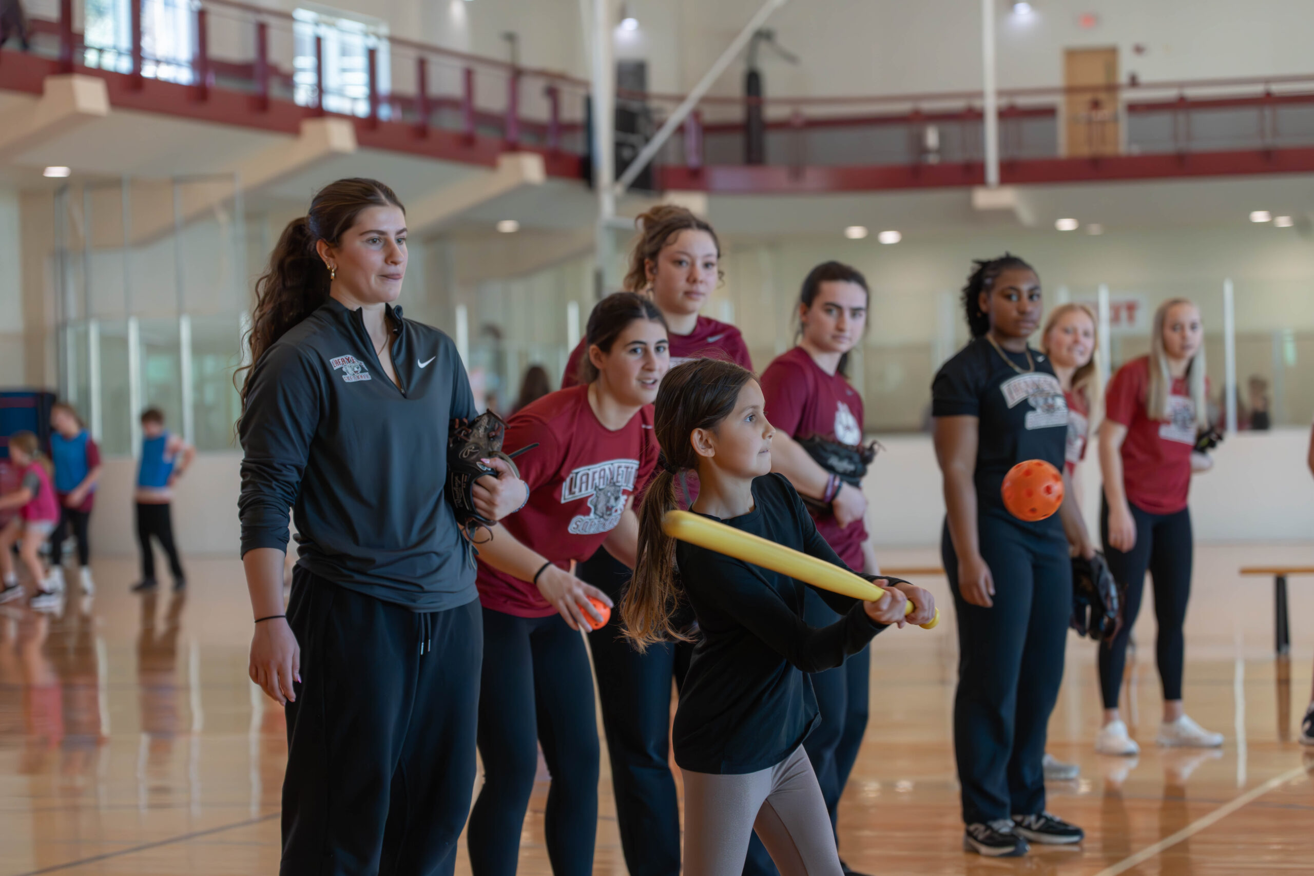 A young girl swings a yellow whiffle ball bat at a red whiffle ball. In the background college aged women athletes can be seen cheering her on. 