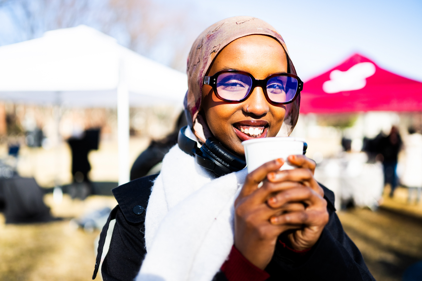 Student drinking hot chocolate during a winter festival. There is a red tent in the background. She is holding a cup of hot chocolate with both hands close to her mouth.