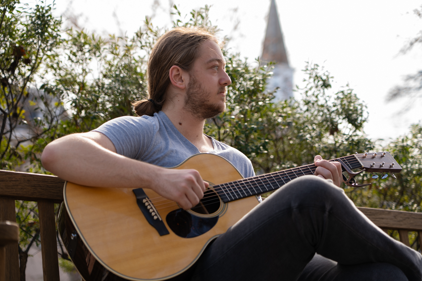 A student sitting and playing an acoustic guitar on a warm day, with bright sunlight. The guitar is brown and the student is wearing a light blue shirt. 