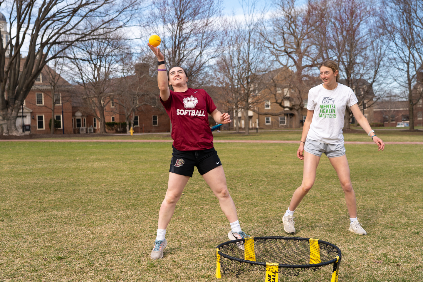 Two students outside at Lafayette College playing Spikeball on a big open grassy field. One student is wearing a maroon t-shirt that says "Softball", she is spiking the ball onto the trampoline. The other student is smiling and laughing. The game is being played under a patchy blue sky.