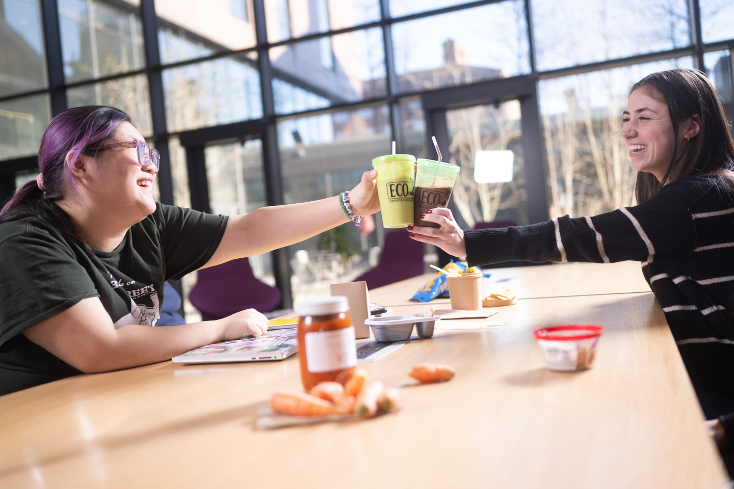 Two students clinking their "Eco Cafe" smoothie cups, smiling and celebrating.