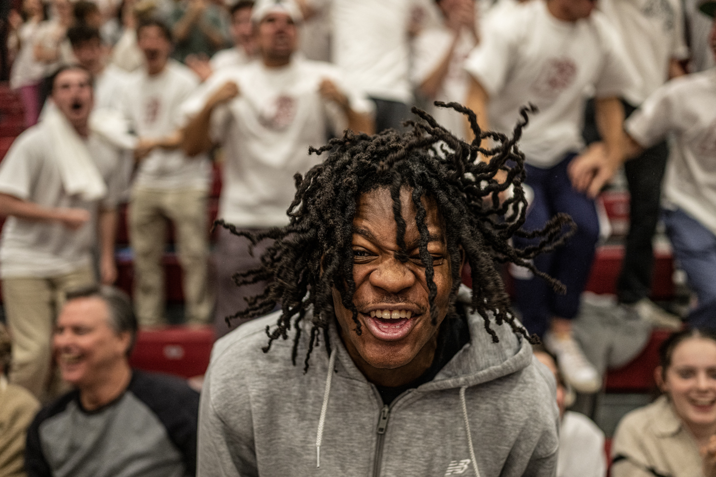 An excited fan cheering wildly at a basketball game. He is wearing a gray hoodie and is surrounded by the student section.