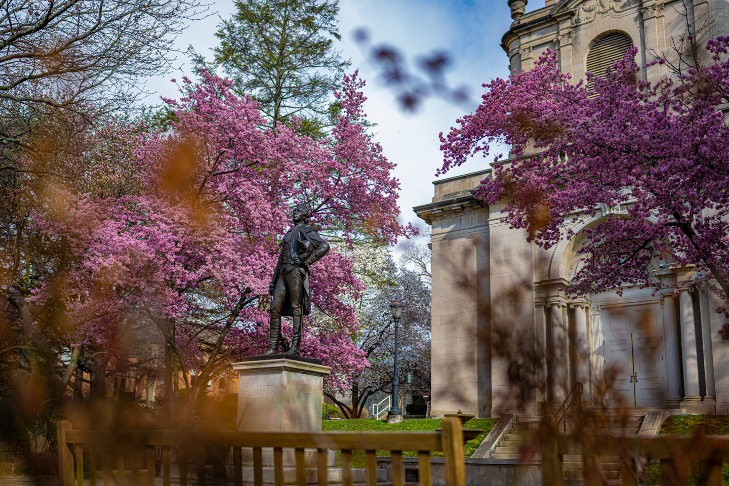 The Marquis statue at Lafayette College surrounded by an abundance of blooming flowers and trees, with a clear blue sky.