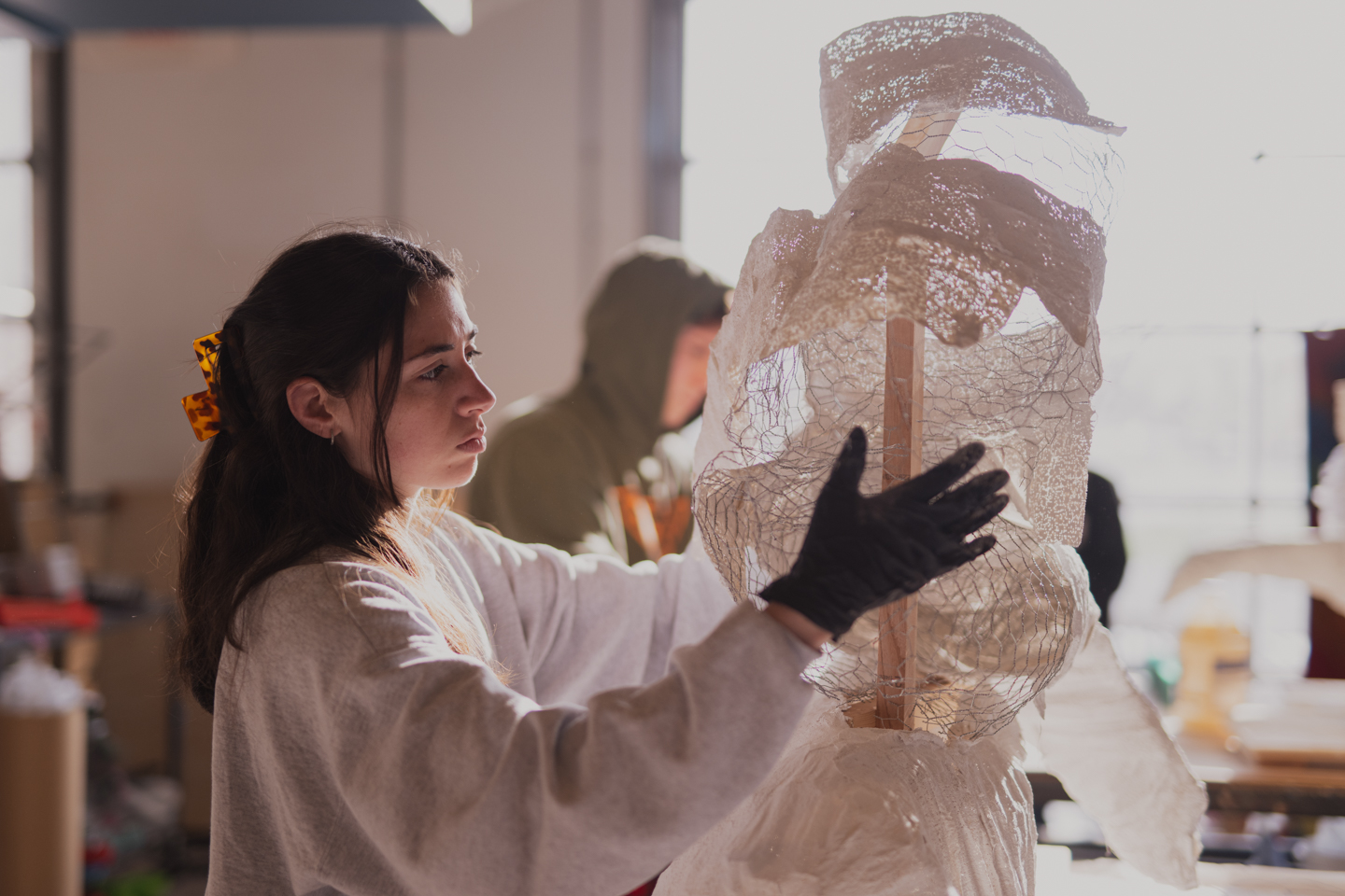 A student calmly working on a sculpture. The room is chaotic filled with various artwork, tools, and materials.