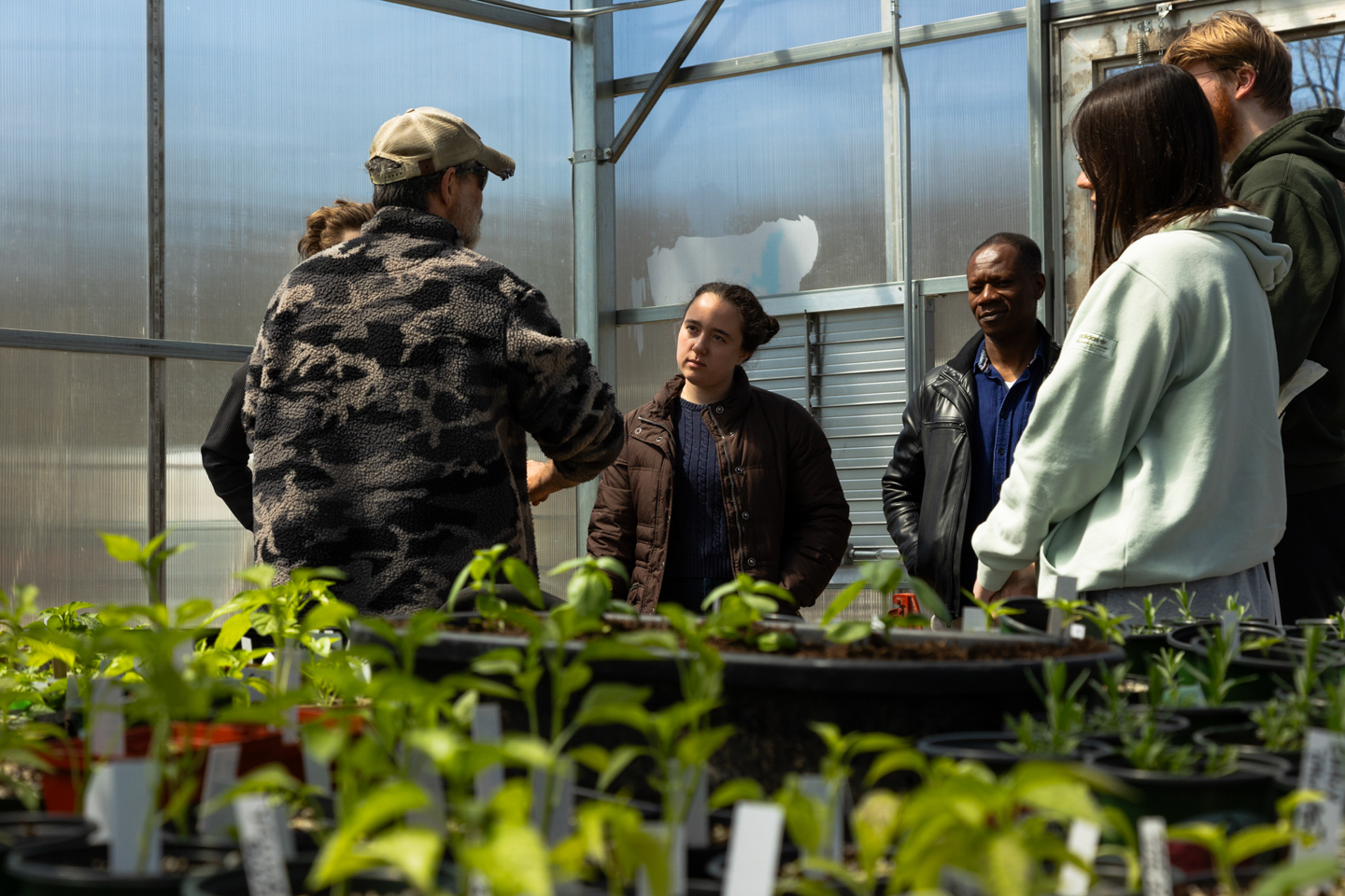 Professor Ernest Nkansah-Dwamena and his class standing in a greenhouse at the Lower Hackett Community Garden, listening to Mark Reid as he gives them a tour.