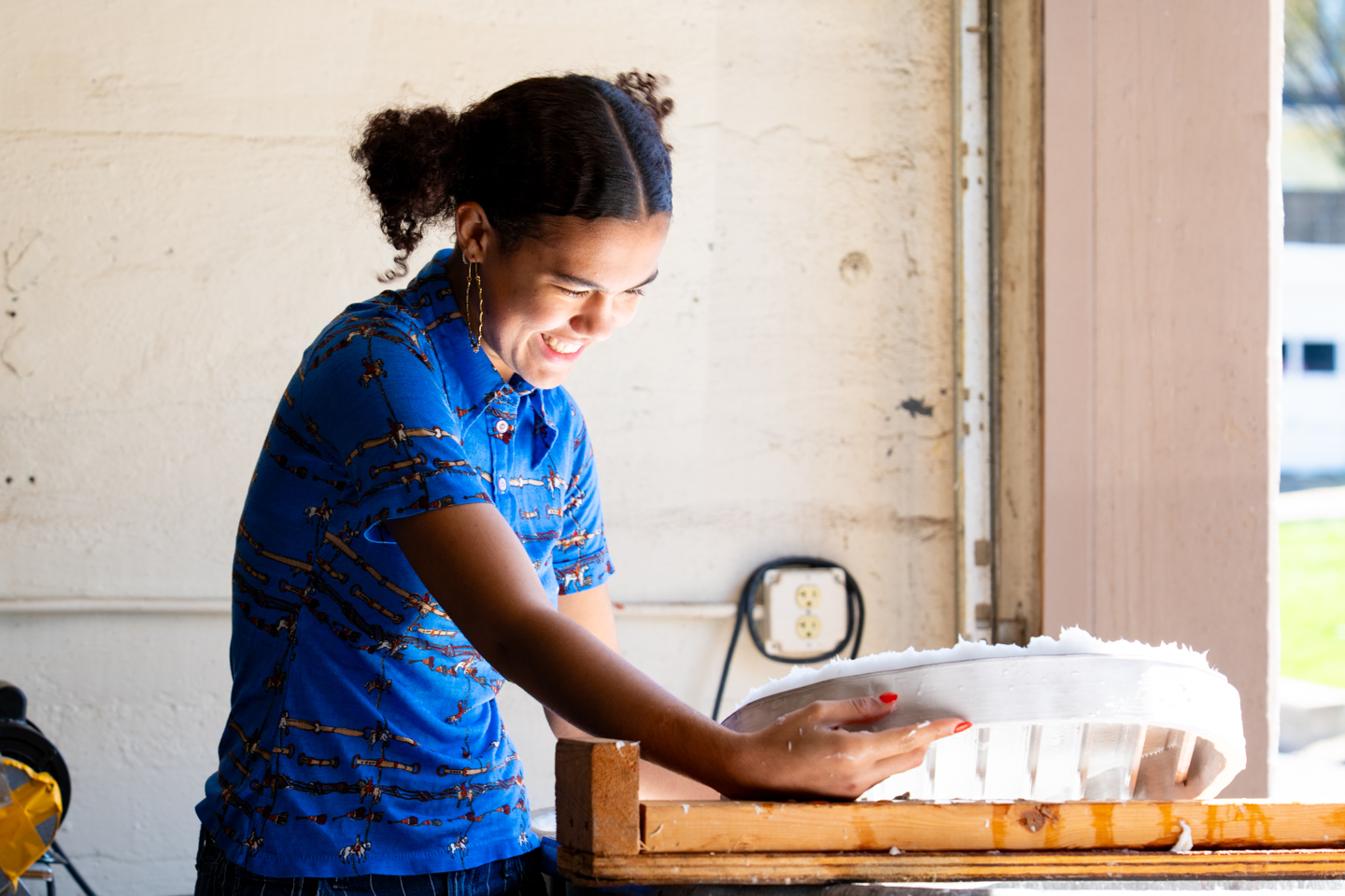 A student in a printmaking class smiles while holding a heart-shaped mold. She is wearing a blue shirt.