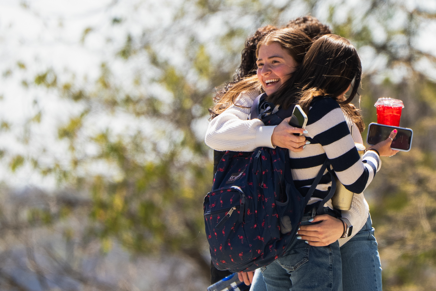 Two students are hugging each other. One of the students is wearing a striped shirt while holding a cell phone and a red drink. It is a bright sunny day. 