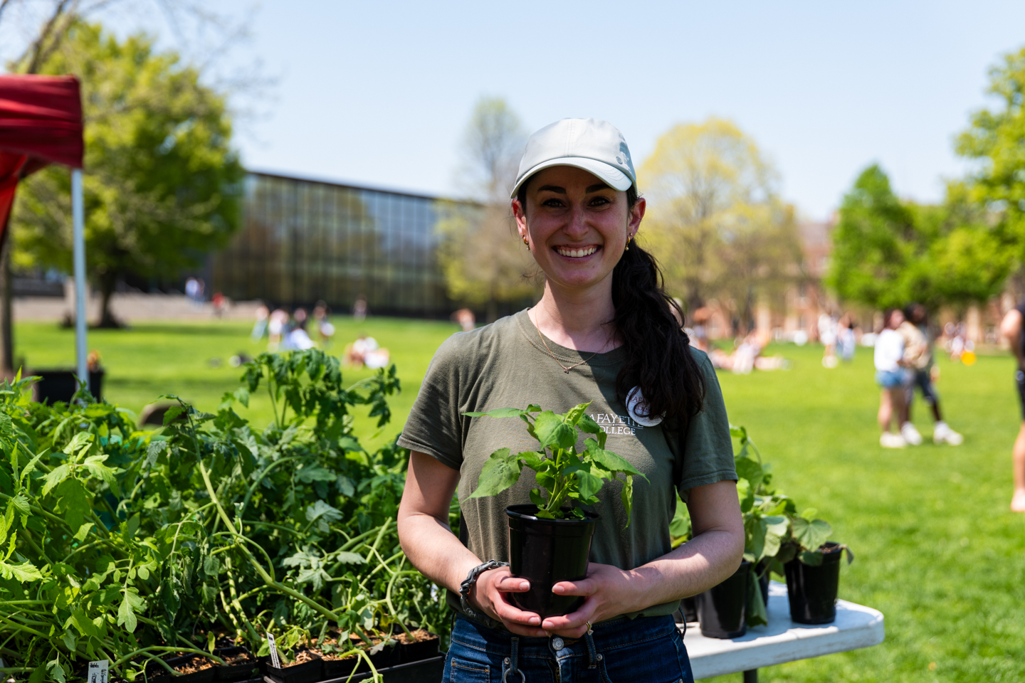 A girl is standing and smiling while holding a plant. There are also various plants for sale behind her. It is a sunny day with a blue sky. In the background, students are gathering in the big grassy field
