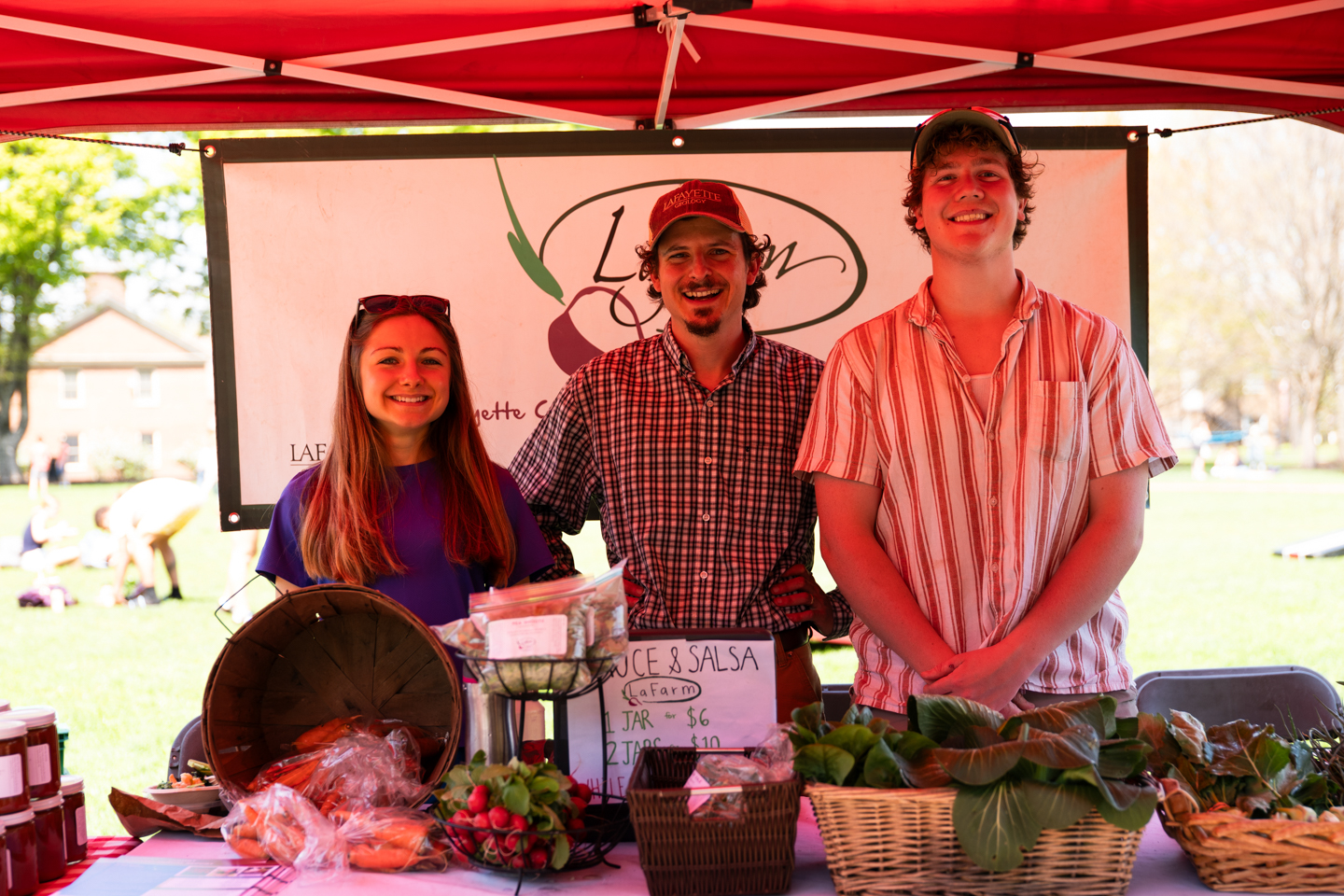 A group picture of 3 people standing under a red tent. In front of them is different kinds of produce for sale.