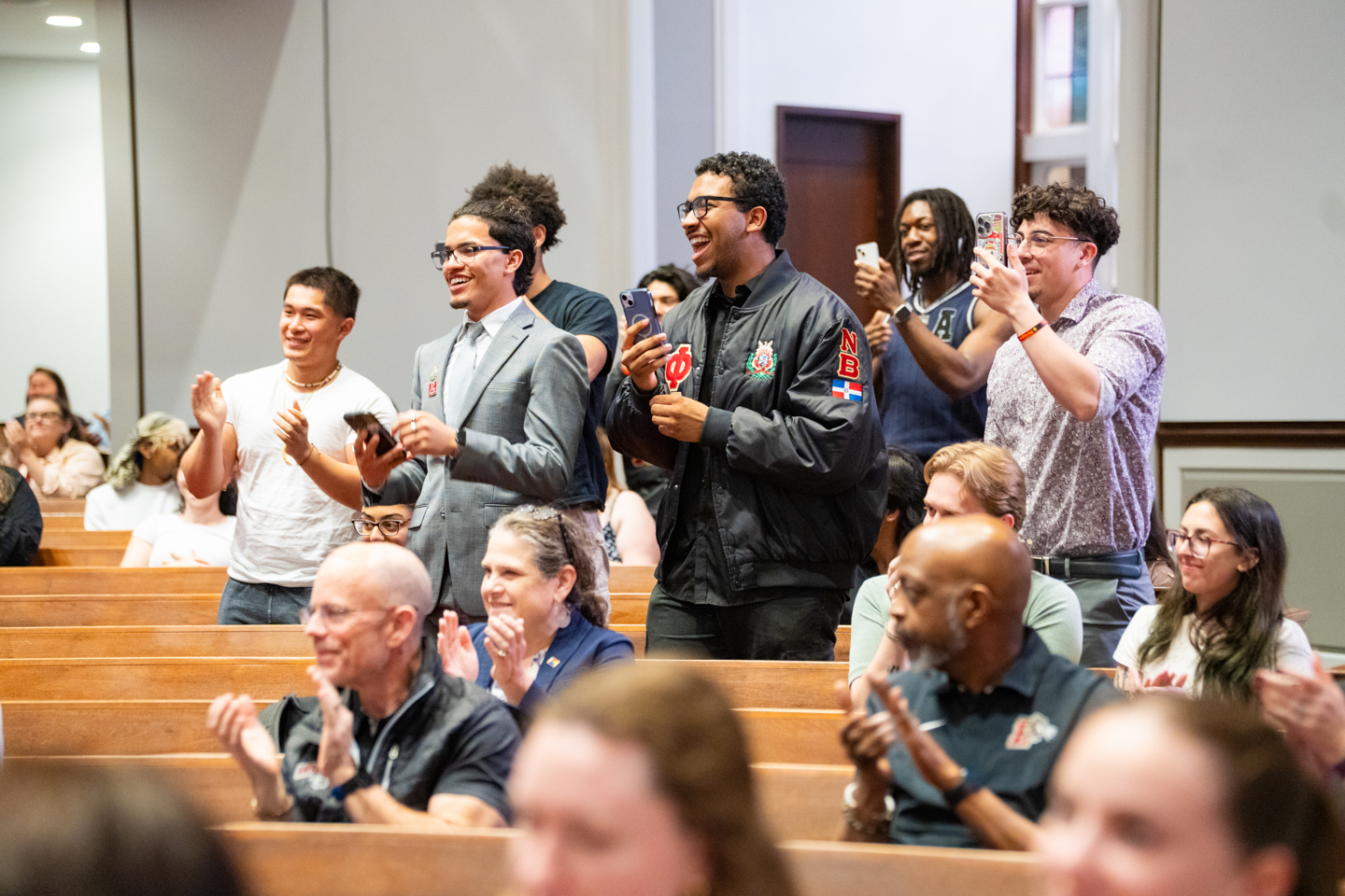 Students standing and cheering enthusiastically during a ceremony as their friend receives an award. 