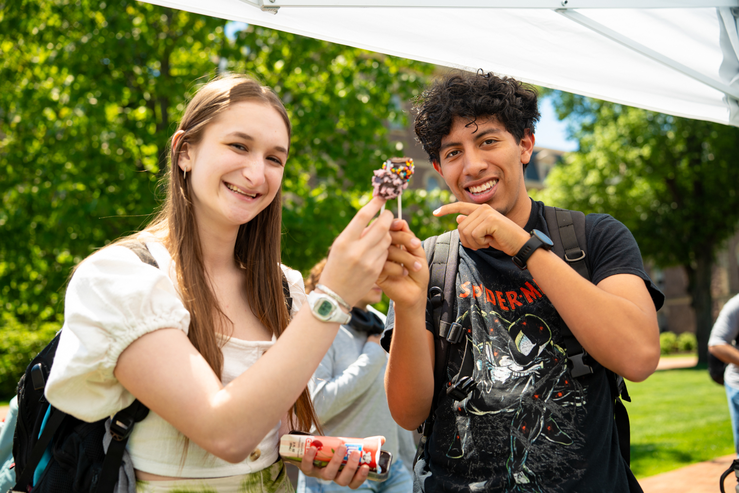 Two students are smiling and toasting cake pops together under a tent during a beautiful spring day. 