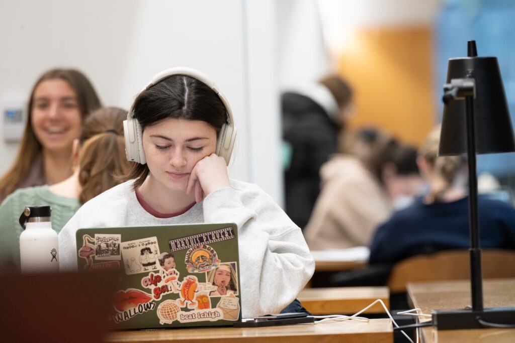 A student has headphones on styudying in front of a computer as other students work in the background