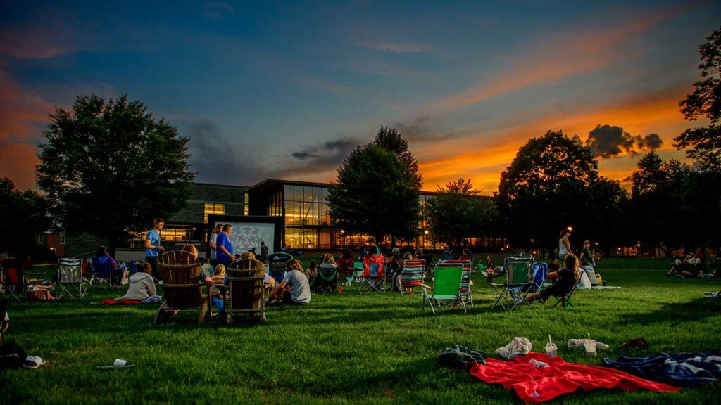 The sun sets as students, staff, and neighbors gather in lawn chairs on the Quad to watch a movie on an inflatable screen