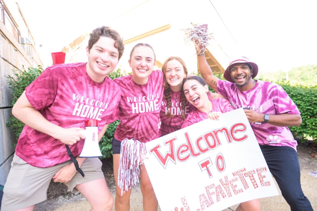 5 Lafayette team members wear maroon tie dye shirts and hold Welcome to Lafayette signs.