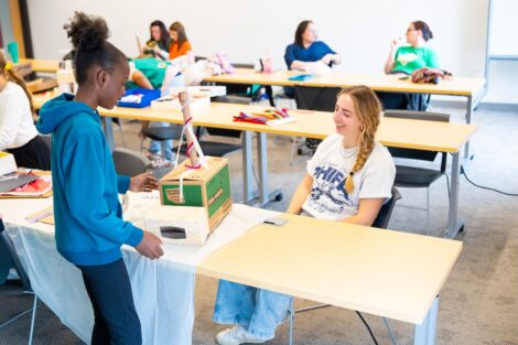 A Lafayette volunteer sits at a table as she helps a Girl Scout construct a rover out of recycled materials.