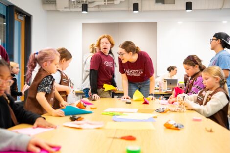 Two SWE volunteers in Lafayette shirts are centered at a table working with a group of Girl Scouts building paper airplanes.
